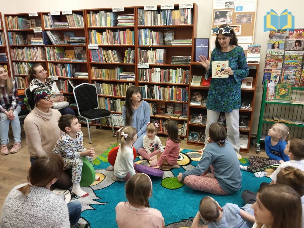 Zdjęcie: Warsztaty dla rodzin w bibliotece na temat czytania wrażeniowego. Dzieci wraz z rodzicami siedzą na niebieskim dywanie i słuchają wypowiedzi zaproszonej pisarki.